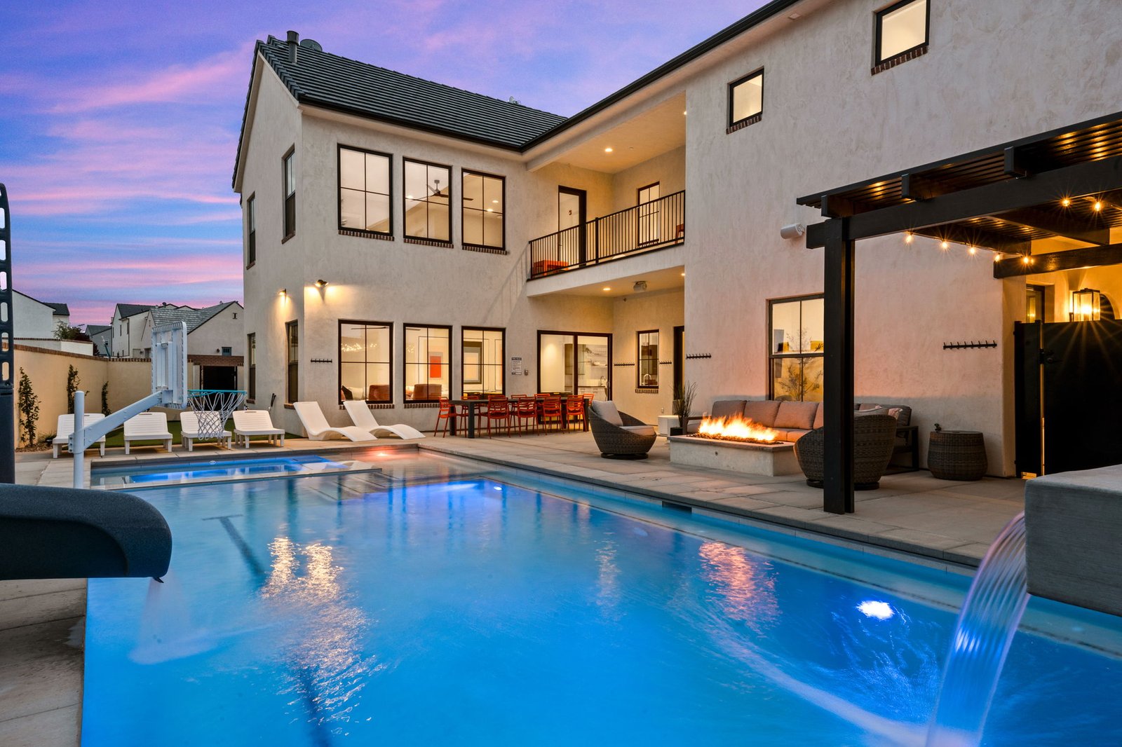 Color House backyard at dusk: glowing pool, waterfall, fire pit lounge, loungers and basketball hoop below the lit-up house